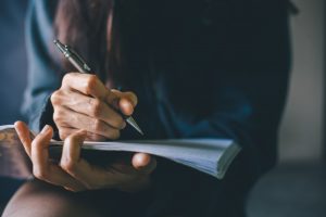 Dark-haired woman writing in notebook that is resting in her lap