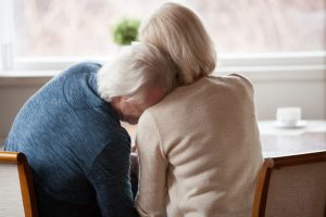 Older man and woman sitting at table grieving, man's head on woman's shoulder