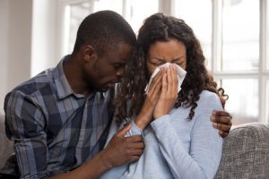 Man and woman sitting on couch, woman crying and blowing nose while husband hugs her in comfort