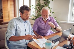 Father and adult son sitting at desk together, looking at documents with computer nearby