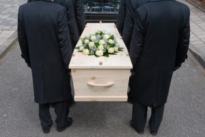 Pallbearers carrying light-colored casket with funeral spray with white roses on top