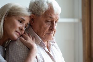 Mature woman standing behind mature man, both sad, her head resting on his shoulder