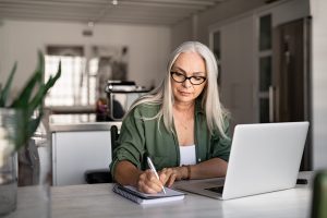 Mature woman sitting at kitchen table with laptop in front of her, writing on notepad