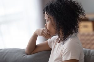 Pensive young woman sitting on couch, hand covering mouth, anxious