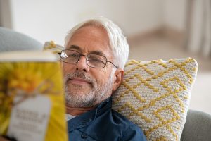 Middle-aged man laying on couch, resting on yellow checkered pillow, reading a book