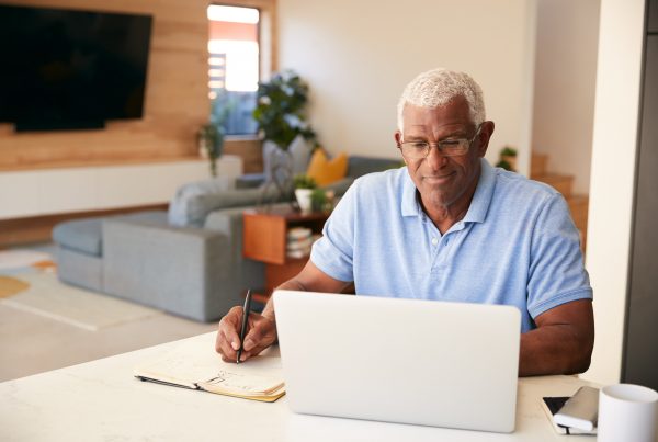 Older man sitting at computer, writing notes in notebook