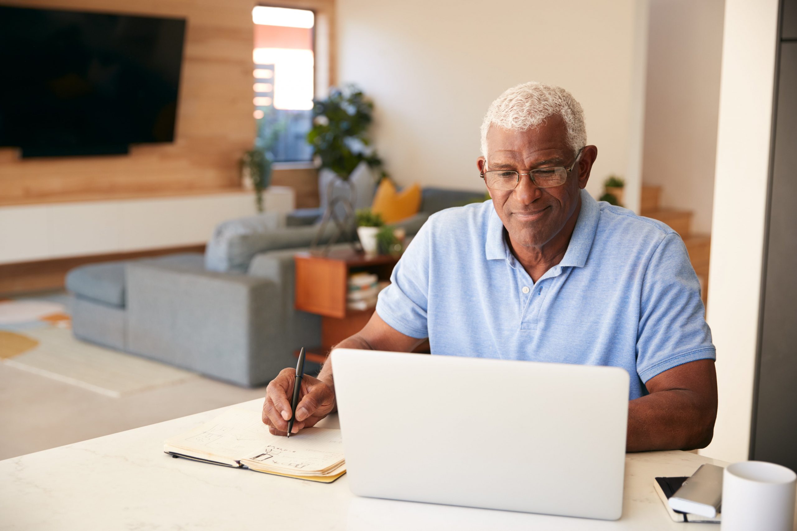 Older man sitting at computer, writing notes in notebook