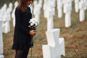 Woman wearing black as she stands next to grave marker and holds white flowers