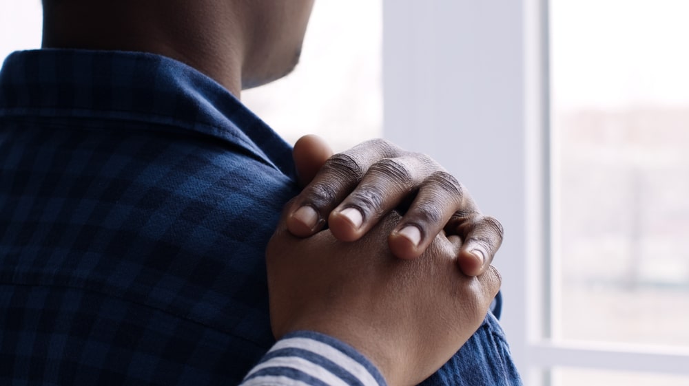 Man looking out window, friend places comforting hand on shoulder