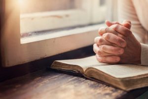 Woman sitting near window with hands clasped on book