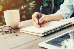 Person sitting at table outside, writing in notebook, expressing feelings