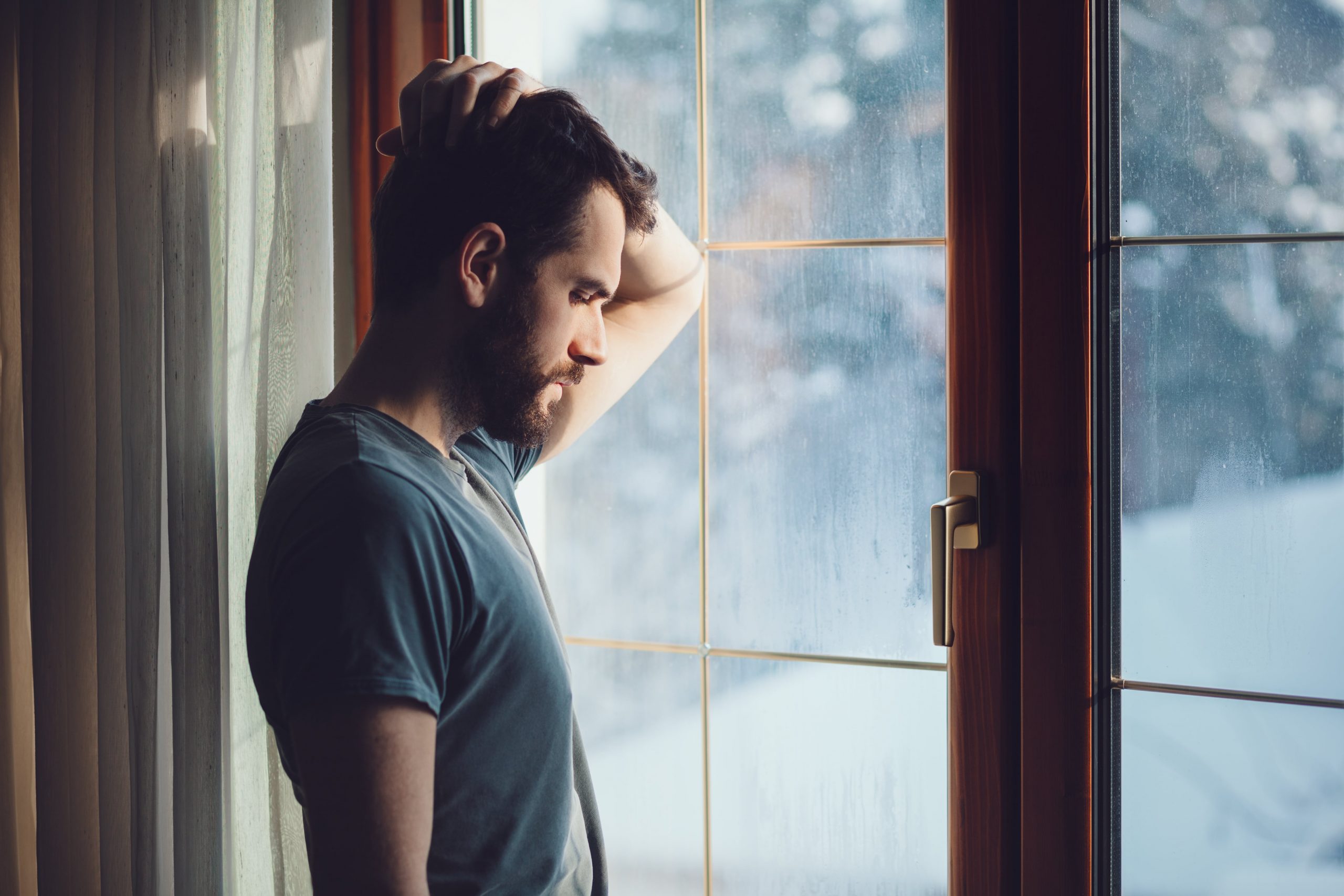 Shadowed man looking out window, hand on head, regretful