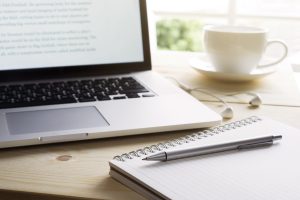 Laptop sitting on desk in natural light with mug, notepad, and pen nearby