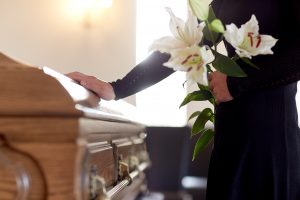 Woman standing next to casket, one hand touching top of casket and the other hand holding white lilies