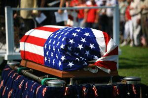 Flag-draped casket sitting on raised area, mourners standing nearby