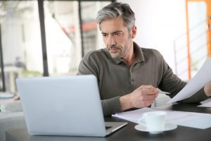 Middle-aged man sitting at home, looking at digital accounts in laptop as he reviews paperwork