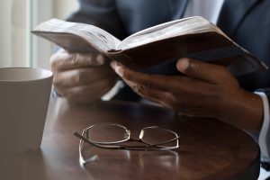 Man sitting at table with mug and glasses, holding and reading Bible