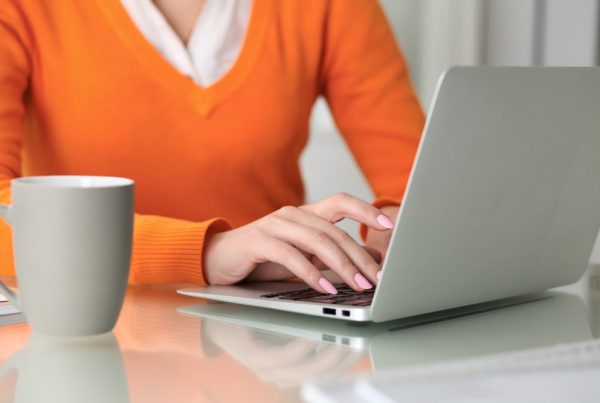 Woman wearing orange sweater sitting at table with laptop, checking on her digital accounts