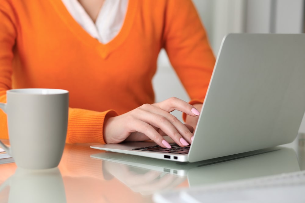 Woman wearing orange sweater sitting at table with laptop, checking on her digital accounts