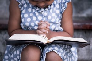 Young girl sitting on steps outside, Bible in lap, hands clasped in prayer