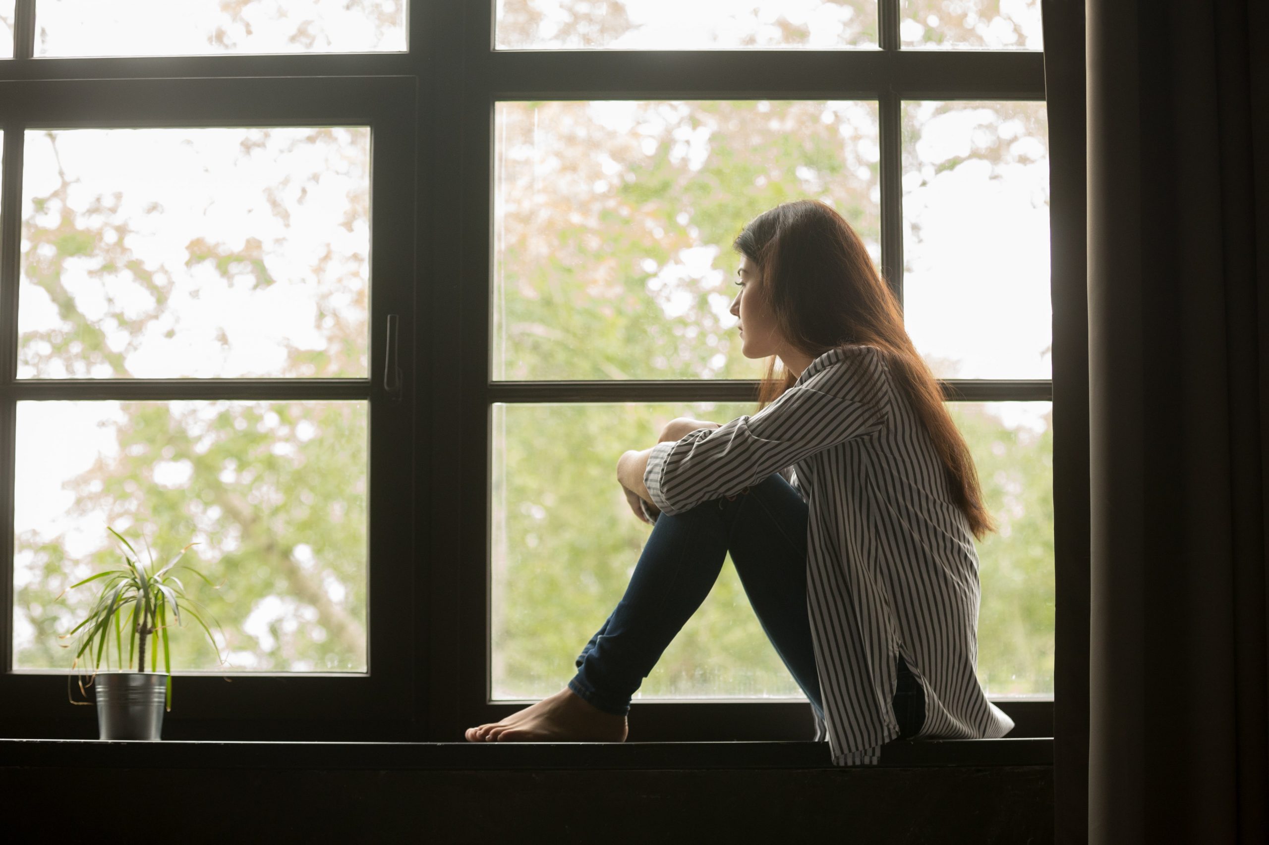 Young woman sitting by window, looking out and thinking quietly