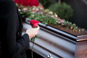 Woman wearing black and holding red rose as she stands beside a dark wood casket covered in flowers