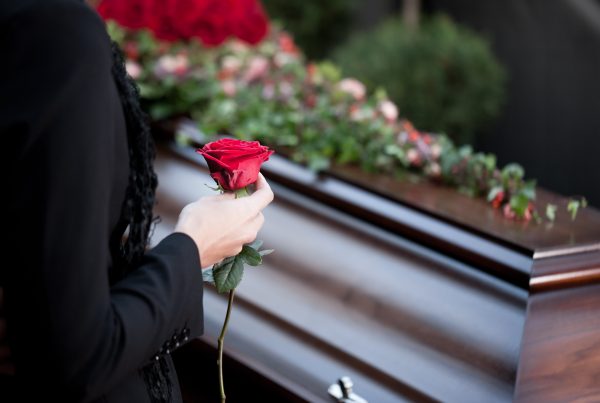 Woman wearing black and holding red rose as she stands beside a dark wood casket covered in flowers