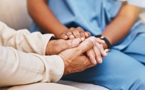 Nurse and hospice patient sitting on couch together, nurse holding patient's hands comfortingly