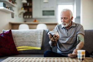 Bearded older man sitting on couch at home, learning how to use blood pressure cuff